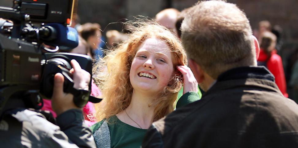 Das Bild zeigt ein blondes Mächen, das vor einer Fernsehkamera von einem Mann interviewt wird.