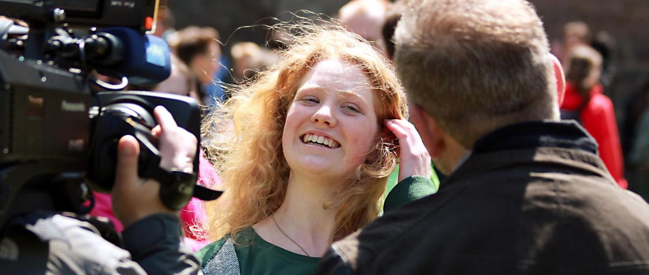 Das Bild zeigt ein blondes Mächen, das vor einer Fernsehkamera von einem Mann interviewt wird.