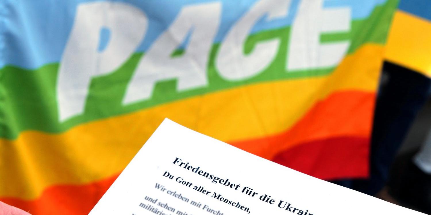 Das Bild zeigt die regenbogenfarbene Friedensflagge mit dem Wort 'pace' und einem davorgehaltenen Blatt Papier mit einem Friedensgebet für die Ukraine