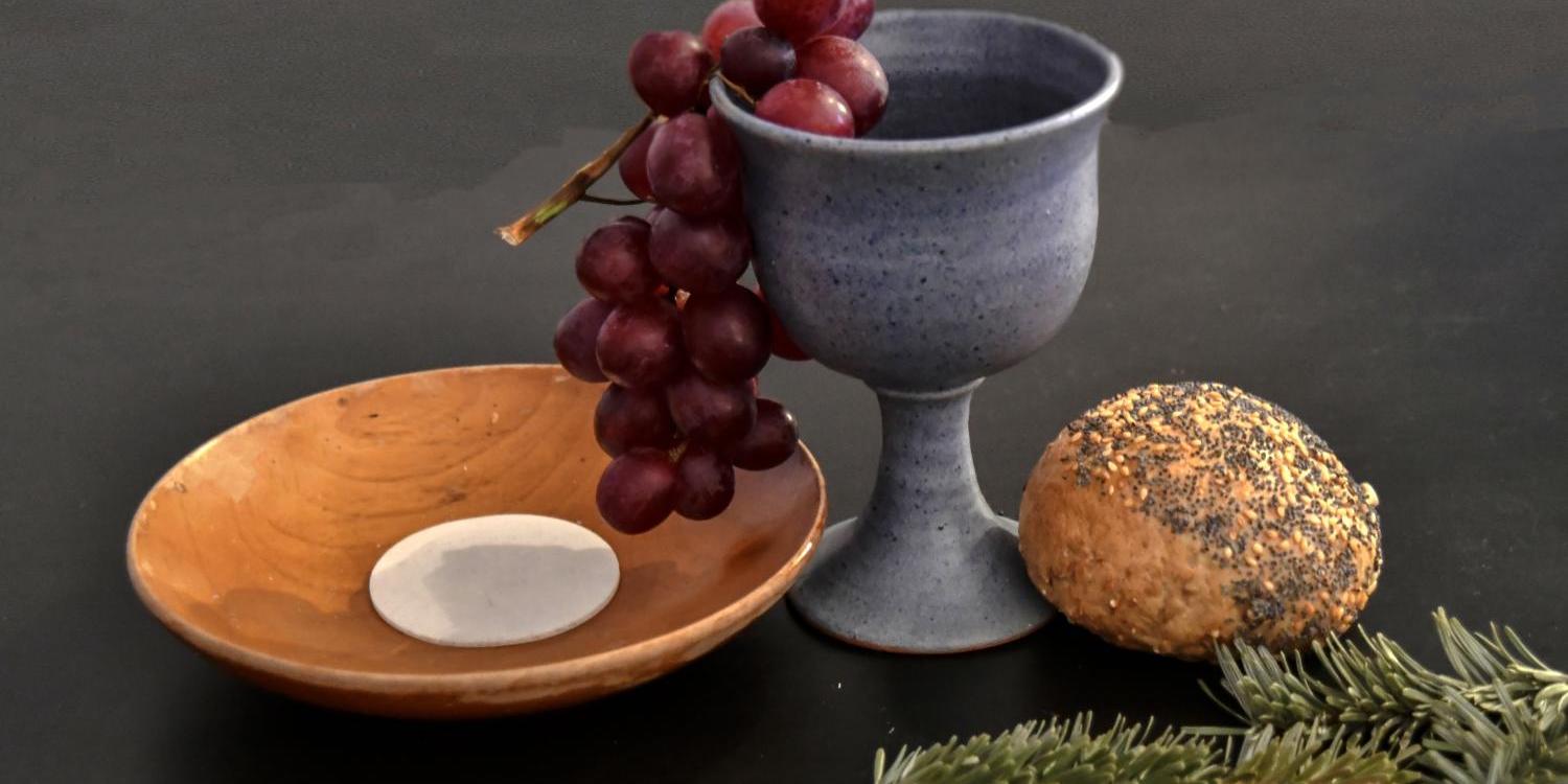 Das Bild zeigt eine Holzschale mit einer Hostie und einen tönernen Kelch, aus dem eine Traube herausrankt. Daneben liegt ein Brötchen und ein Tannenzweig.