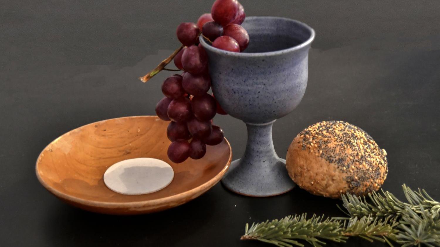 Das Bild zeigt eine Holzschale mit einer Hostie und einen tönernen Kelch, aus dem eine Traube herausrankt. Daneben liegt ein Brötchen und ein Tannenzweig.