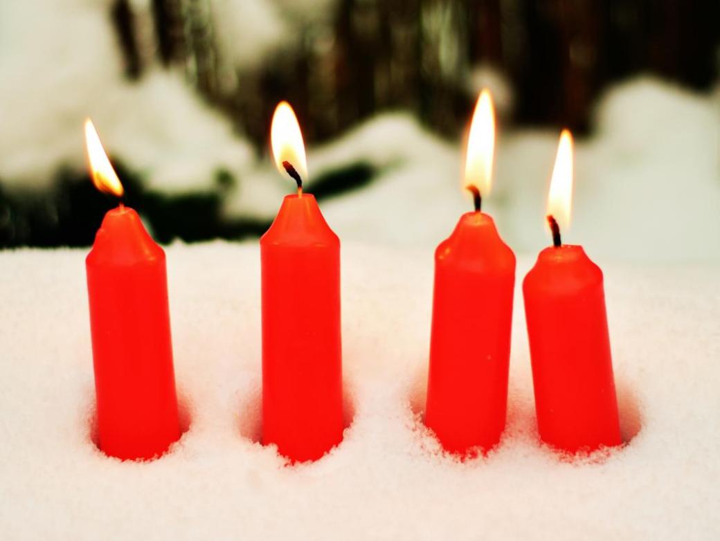 Das Bild zeigt vier rote brennende Kerzen, die im Schnee stecken.