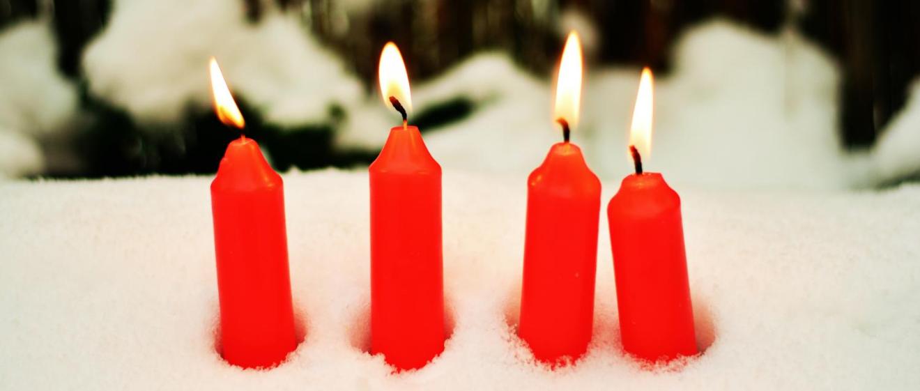 Das Bild zeigt vier rote brennende Kerzen, die im Schnee stecken.