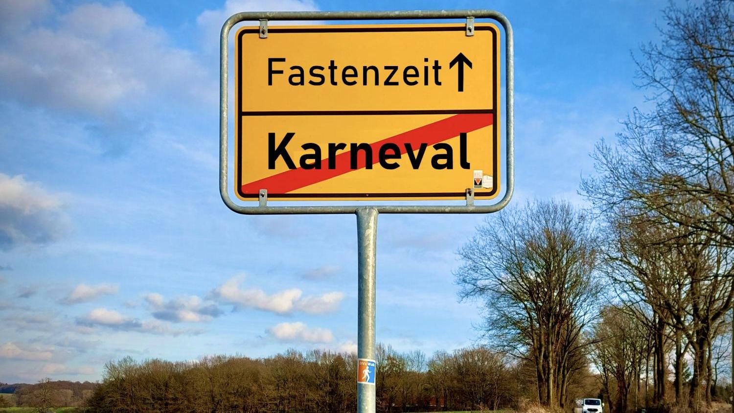 Das Bild zeigt ein Ortsausgangsschild einer Gemeinde, auf dem unten das rot durchgestrichene Wort 'Karneval' steht und obendrüber mit einem Pfeil nach oben das Wort 'Fastenzeit'