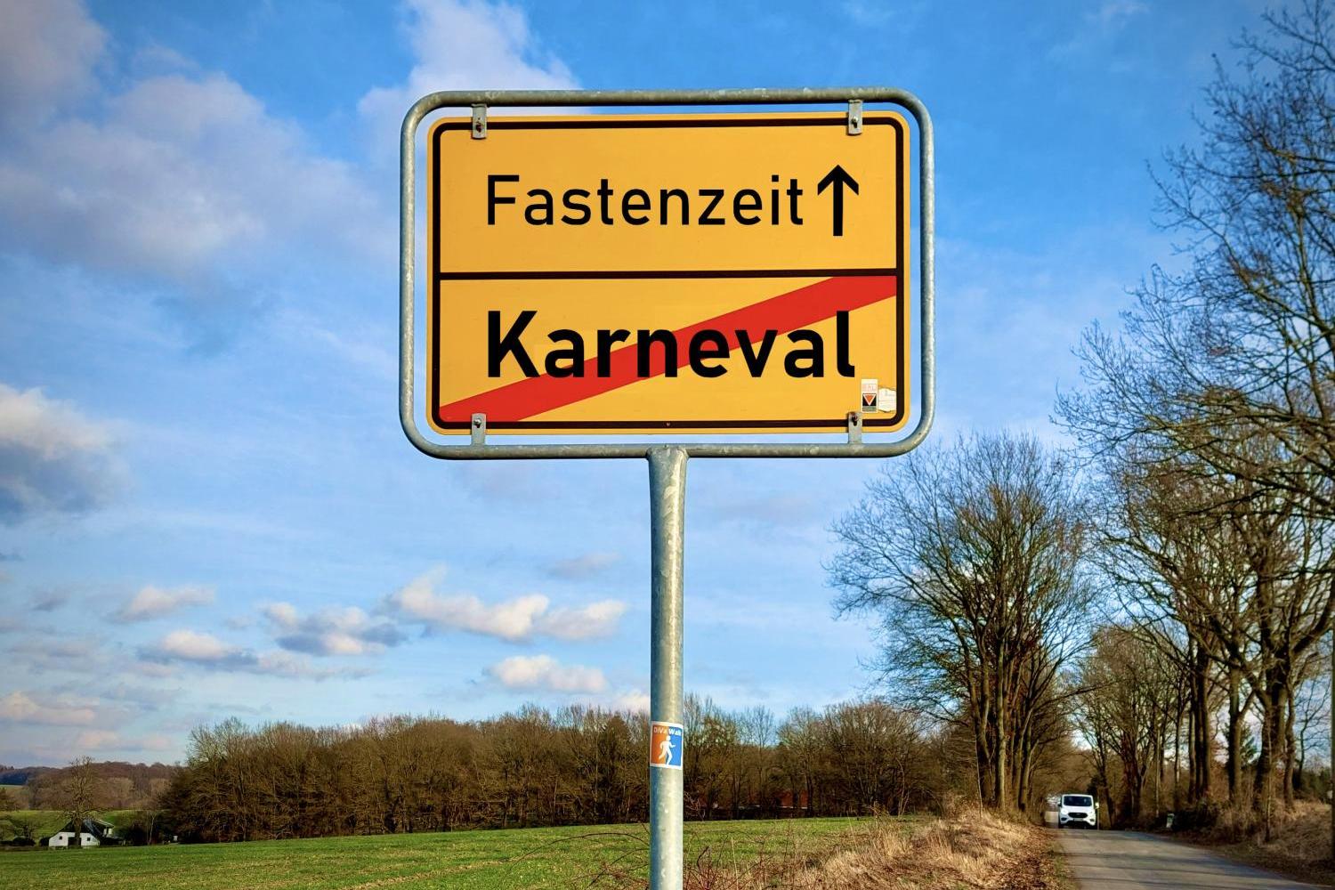 Das Bild zeigt ein Ortsausgangsschild einer Gemeinde, auf dem unten das rot durchgestrichene Wort 'Karneval' steht und obendrüber mit einem Pfeil nach oben das Wort 'Fastenzeit'