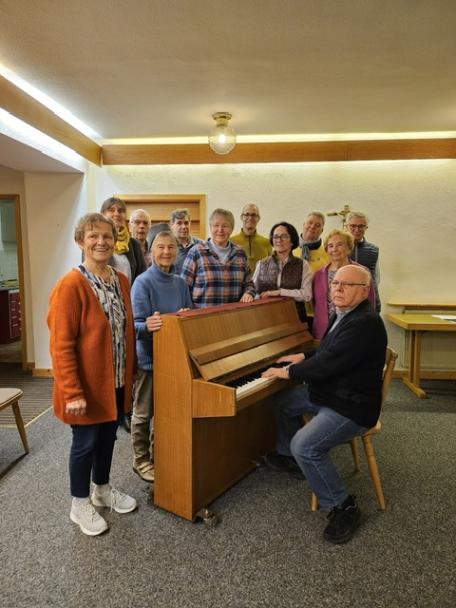 Das Bild zeigt den Kirchenchor Norheim bei einer Probe um ein Klavier stehend. Am Klavier sitzt der Chorleiter