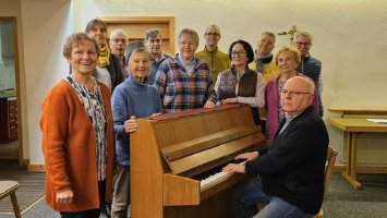 Das Bild zeigt den Kirchenchor Norheim bei einer Probe um ein Klavier stehend. Am Klavier sitzt der Chorleiter