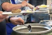 Das Bild zeigt Hände von Personen, die Essen aus einem Warmhaltebehälter auf einen Teller geben.