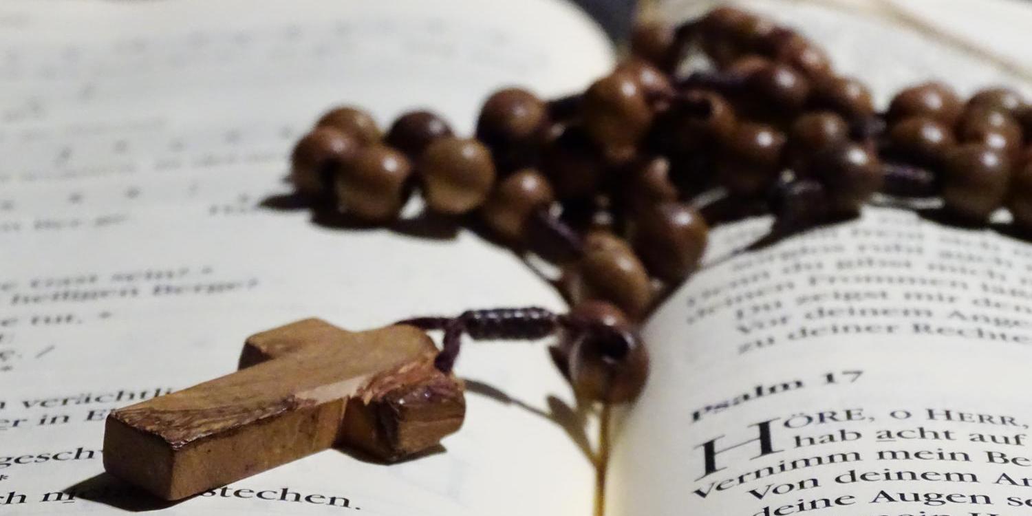 Holzrosenkranz mit Kreuz liegt auf aufgeschlagenem Buch.